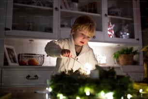 Retrato do menino pequeno dentro de casa no Natal, velas de relâmpago na coroa de flores.