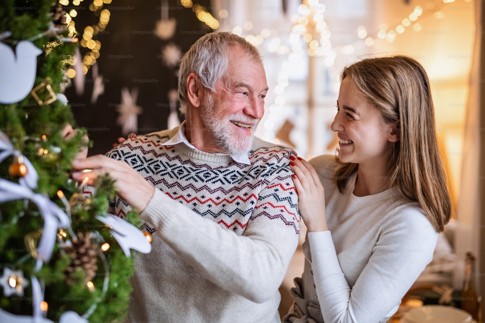 Happy young woman with senior grandfather indoors at home at Christmas, decorating tree.