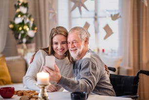 Portrait of young woman with grandfather indoors at home at Christmas, taking selfie with smartphone.