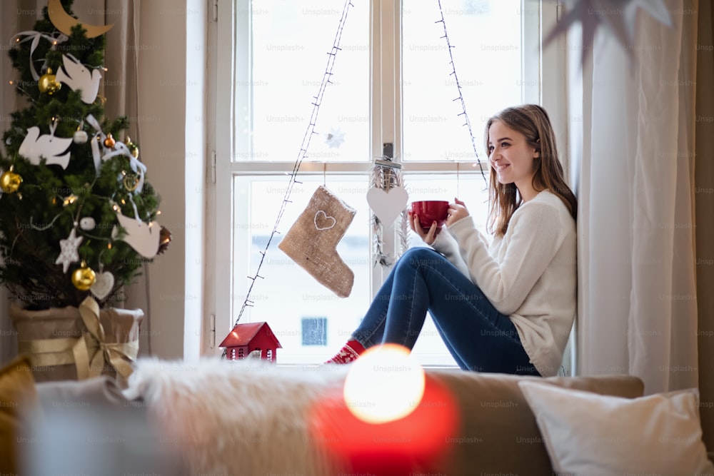 Portrait of young woman indoors at home at Christmas, sitting on window sill with tea.