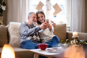 Portrait of young woman with grandfather indoors at home at Christmas, taking selfie.