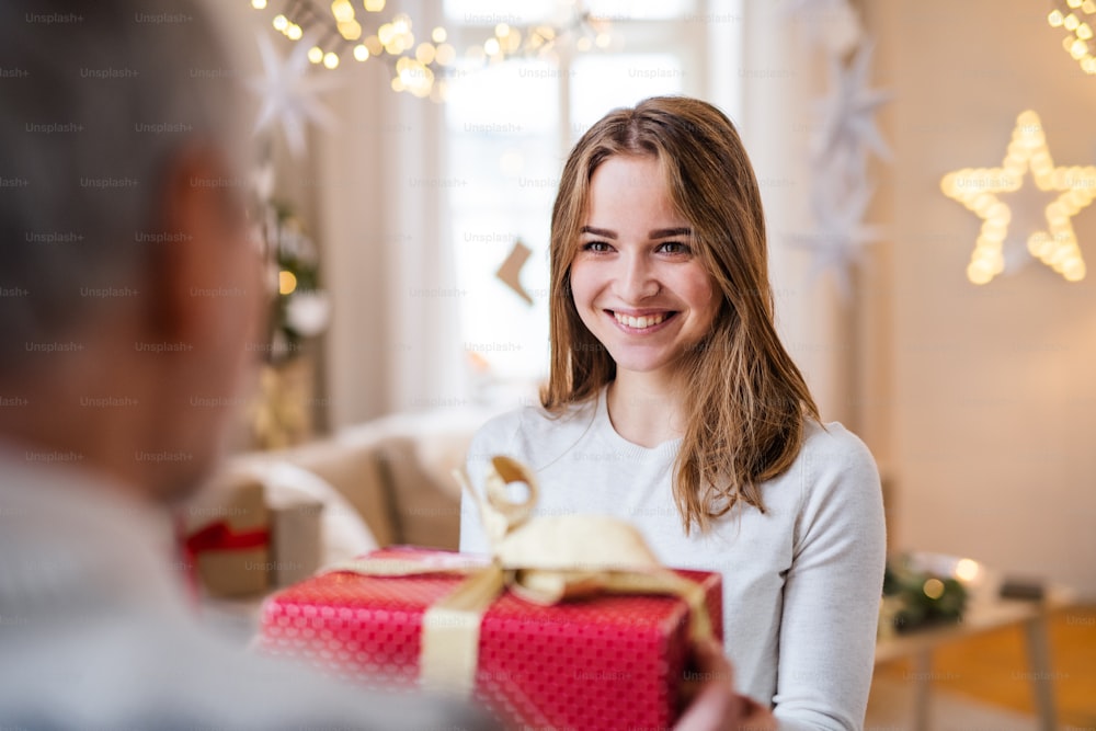 Portrait of young woman giving present to unrecognizable grandfather indoors at home at Christmas.