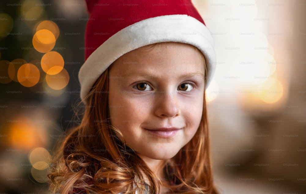 Ritratto ravvicinato di una bambina con il cappello di Babbo Natale in casa a Natale, guardando la macchina fotografica.