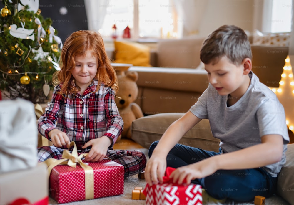 Kleines Mädchen und Junge im Pyjama zu Weihnachten drinnen zu Hause und öffnen morgens Geschenke.