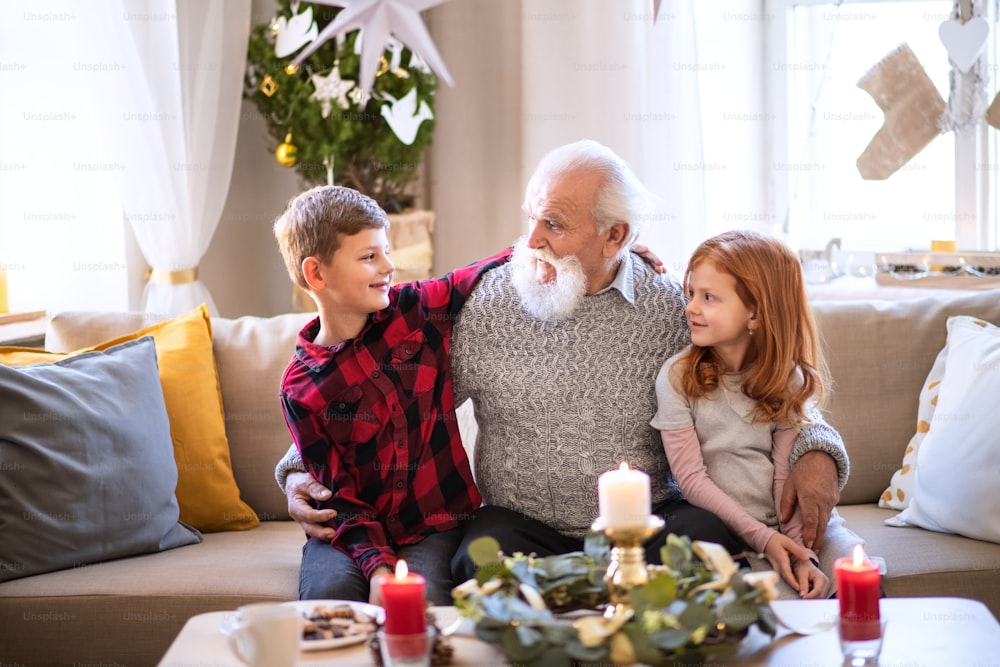 Portrait de jeunes enfants avec grand-père aîné à l’intérieur à la maison à Noël, en train de parler.