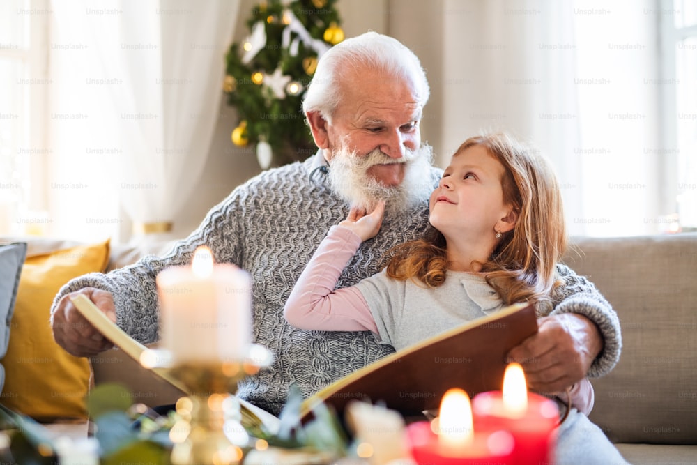 Small girl with senior grandfather indoors at home at Christmas, looking at album with photographs.