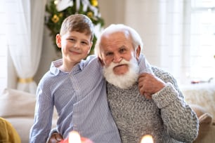 Portait of small boy with senior grandfather indoors at home at Christmas, looking at camera.