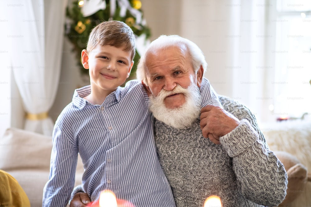 Portait of small boy with senior grandfather indoors at home at Christmas, looking at camera.