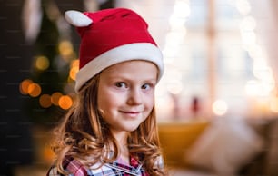 Close-up portrait of small girl with Santa hat indoors at home at Christmas, looking a camera.