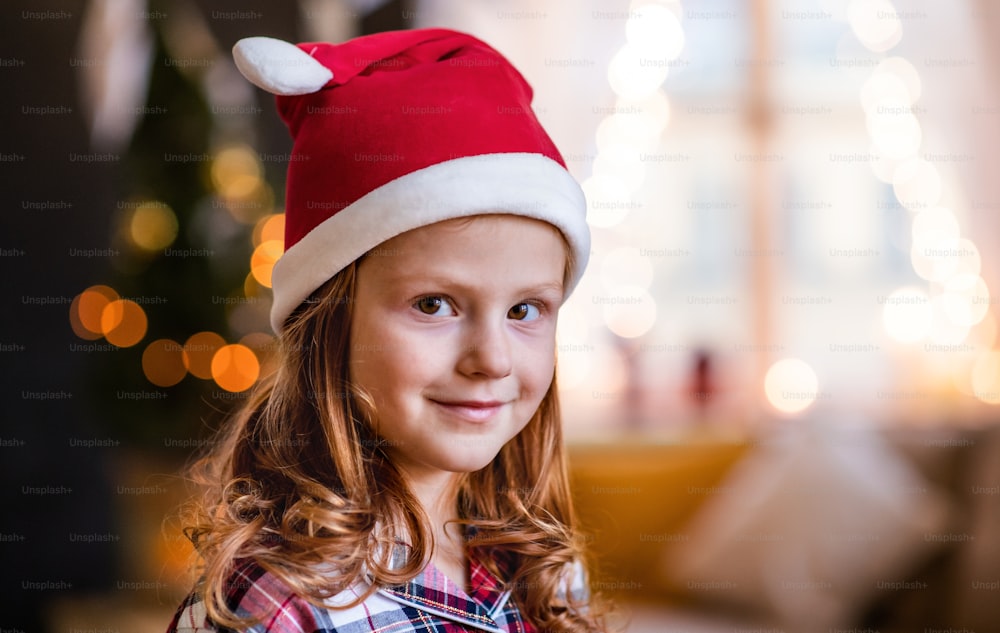 Retrato en primer plano de una niña pequeña con sombrero de Santa en el interior de su casa en Navidad, mirando una cámara.