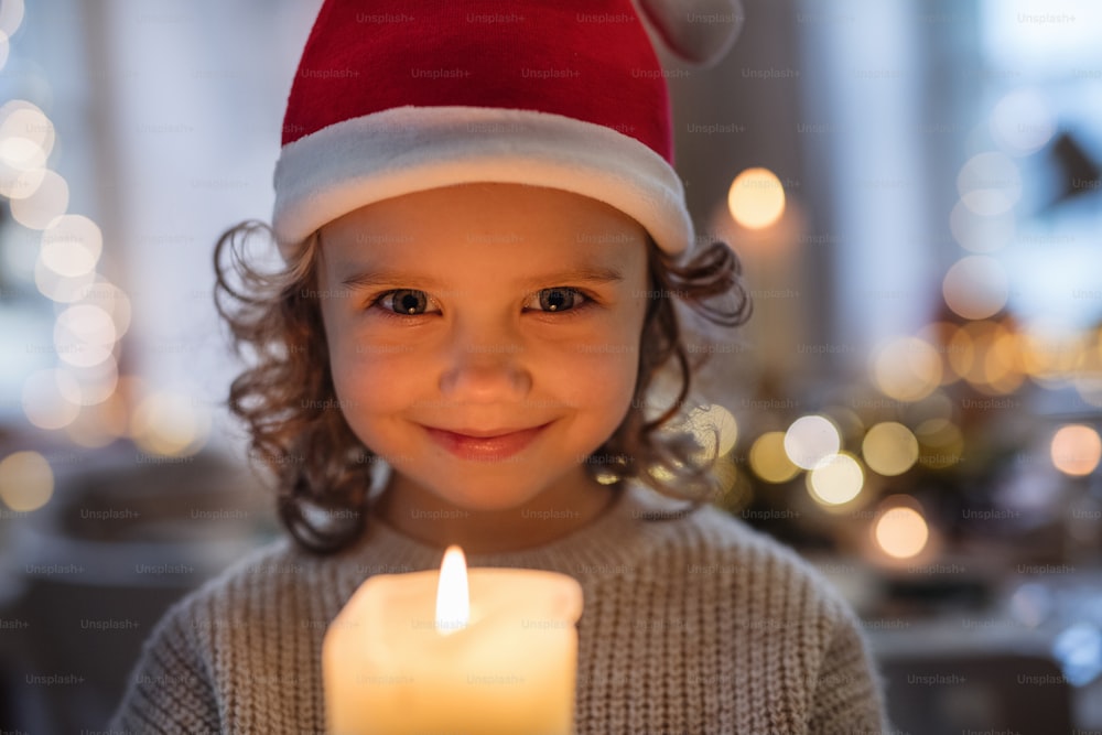 크리스마스에 실내에서 산타 모자를 쓰고 촛불을 들고 있는 쾌활한 어린 소녀의 정면보기.