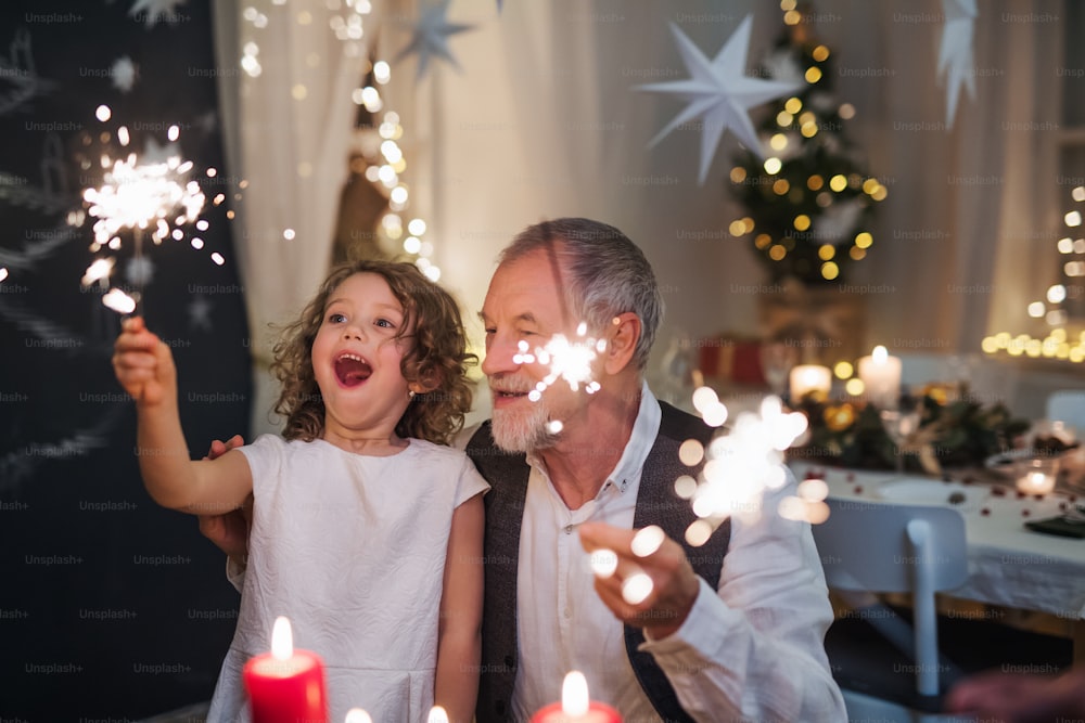 Portrait of senior grandfather with small granddaughter indoors at Christmas, sitting at table with sparklers.