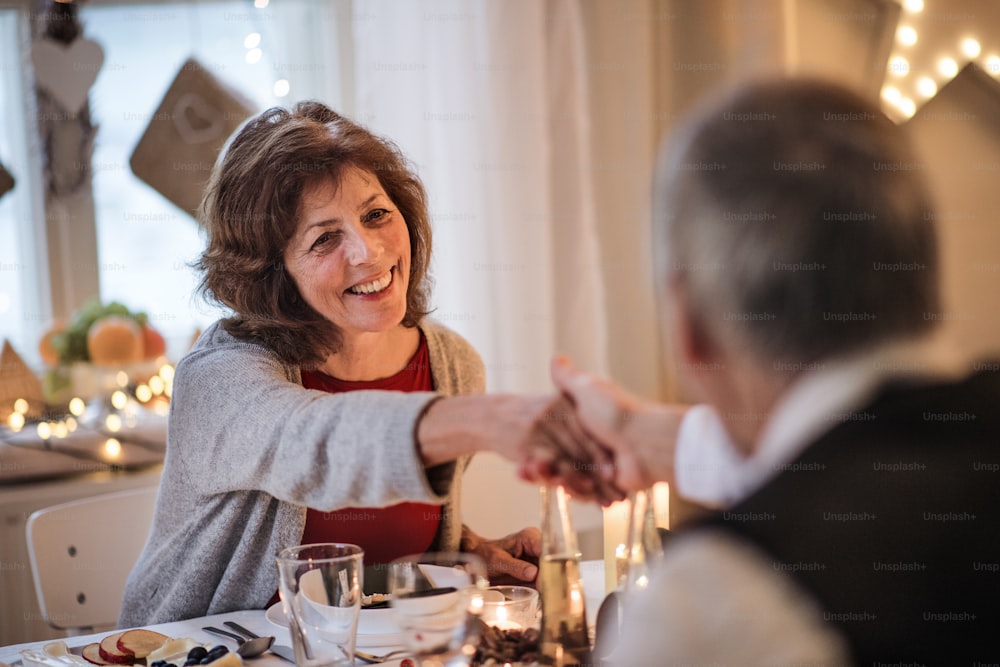 Portrait d’un couple heureux de personnes âgées amoureux à l’intérieur à la maison assis à la table à Noël, en train de parler.
