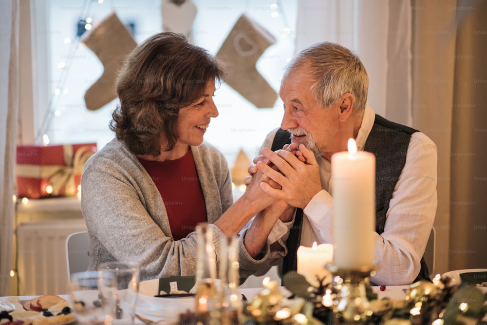Portrait d’un couple de personnes âgées heureux à l’intérieur à la maison assis à la table à Noël, en train de parler.