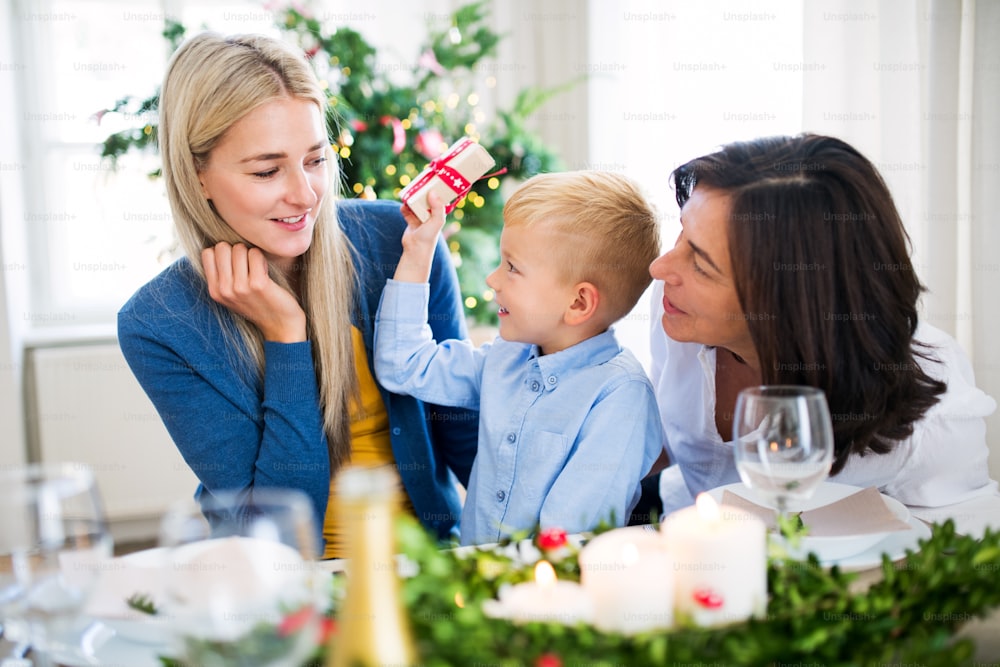 A small boy with present and mother and grandmother sitting at a table at home at Christmas time.