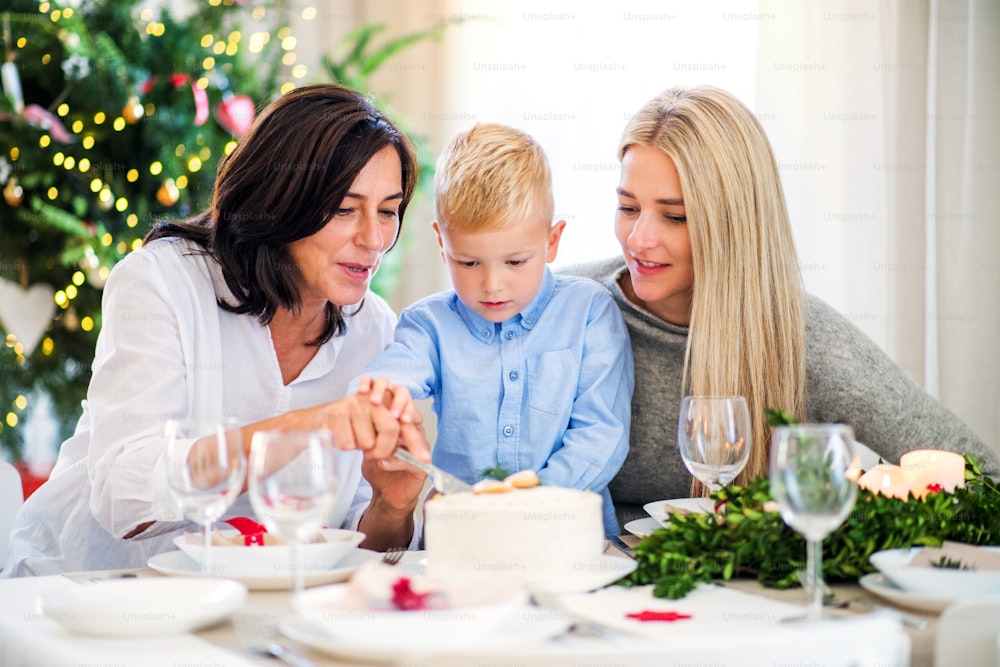 Um menino pequeno com mãe e avó sentadas à mesa, cortando um bolo na época do Natal.