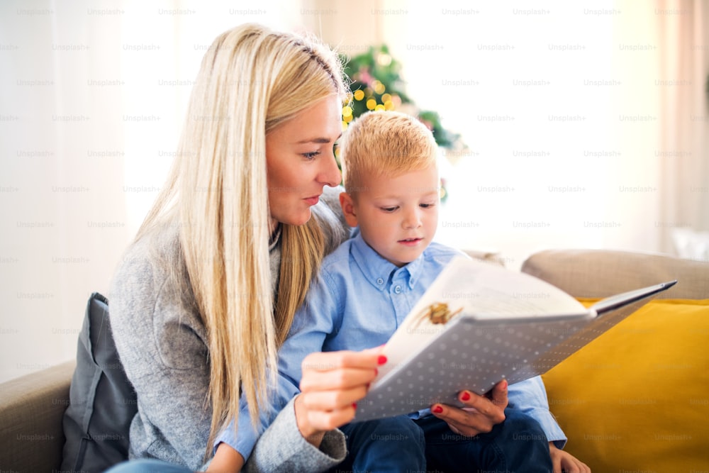 A mother and small boy sitting on a sofa at home at Christmas time, reading a story from a book.