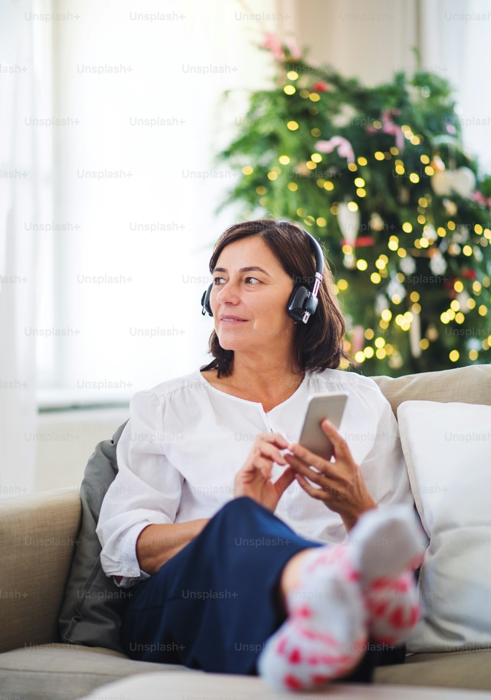 Eine ältere Frau mit Kopfhörern und Smartphone, die zu Hause auf einem Sofa sitzt und zur Weihnachtszeit Musik hört.