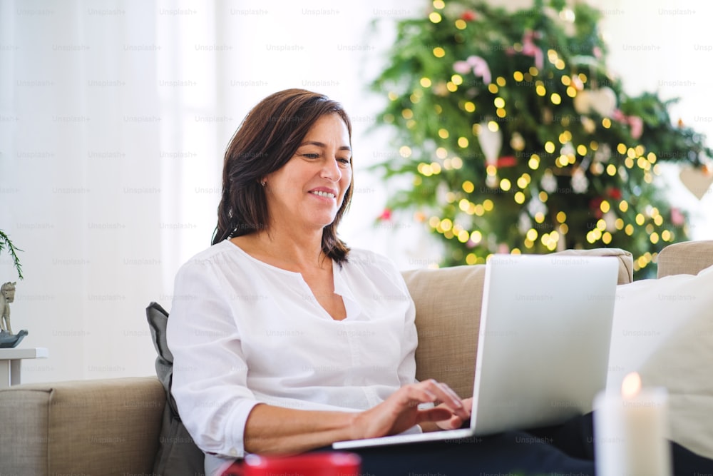 Una donna anziana felice con il computer portatile seduta su un divano a casa nel periodo natalizio.