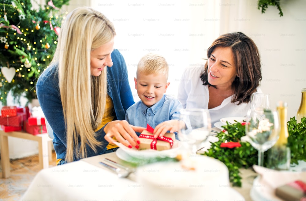 Un bambino con regalo e madre e nonna seduti a un tavolo a casa nel periodo natalizio.