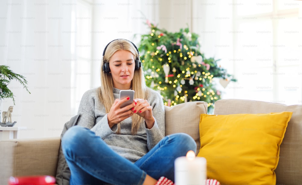 Una giovane donna con cuffie e smartphone seduta su un divano di casa nel periodo natalizio, ascoltando musica.