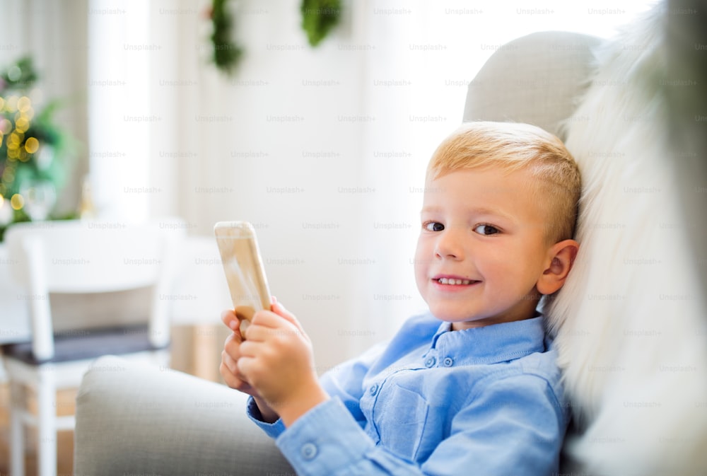Un niño pequeño con un teléfono inteligente sentado en un sillón en casa en Navidad, jugando.