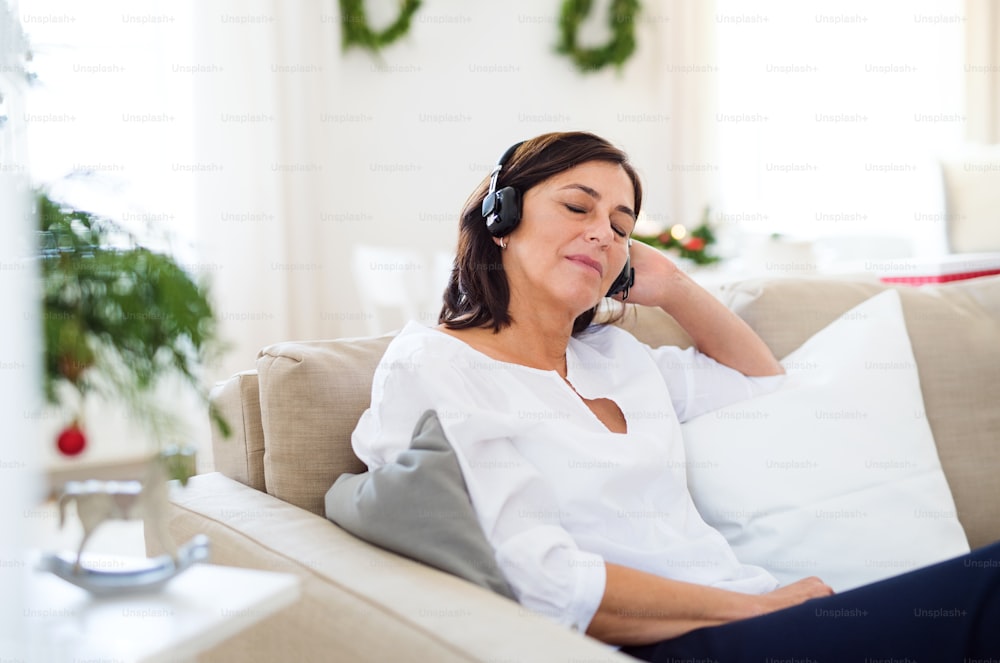 Una mujer mayor con auriculares y ojos cerrados sentada en un sofá en casa, escuchando música en Navidad.