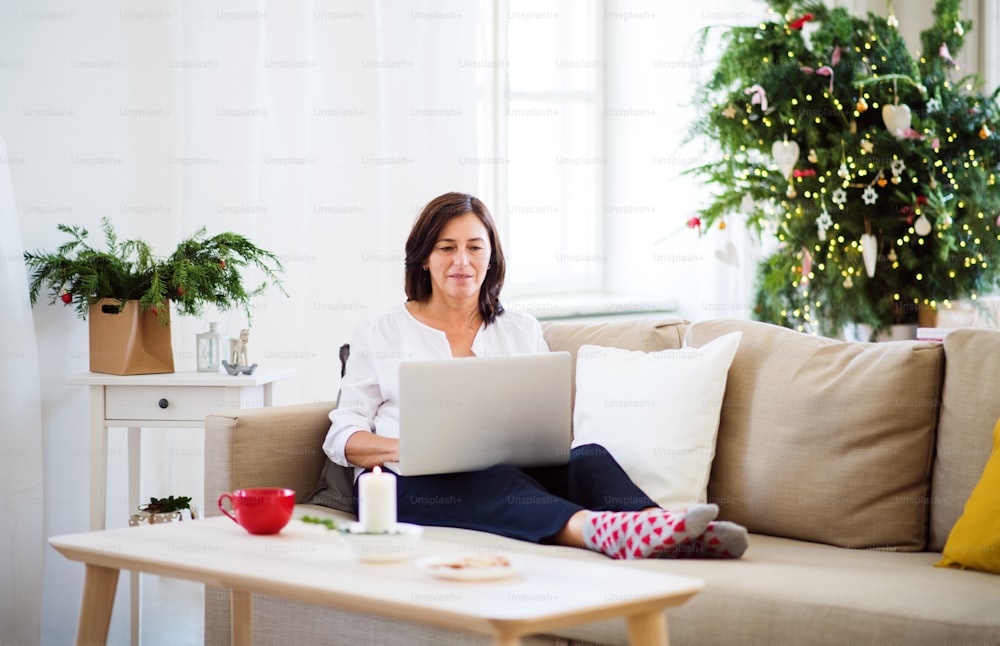 Una mujer mayor feliz con computadora portátil sentada en un sofá en casa en Navidad.