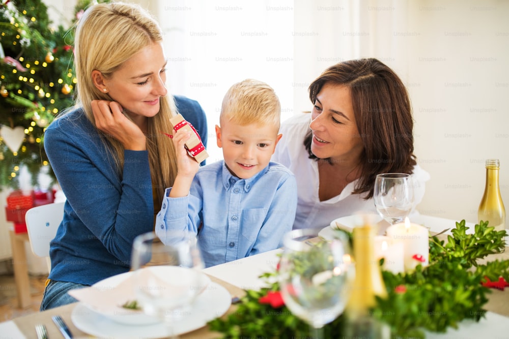 Um menino pequeno com presente e mãe e avó sentados em uma mesa em casa na época do Natal.