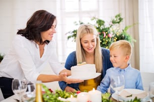Un niño pequeño con madre mirando a la abuela poniendo un pastel en la mesa en casa en Navidad.