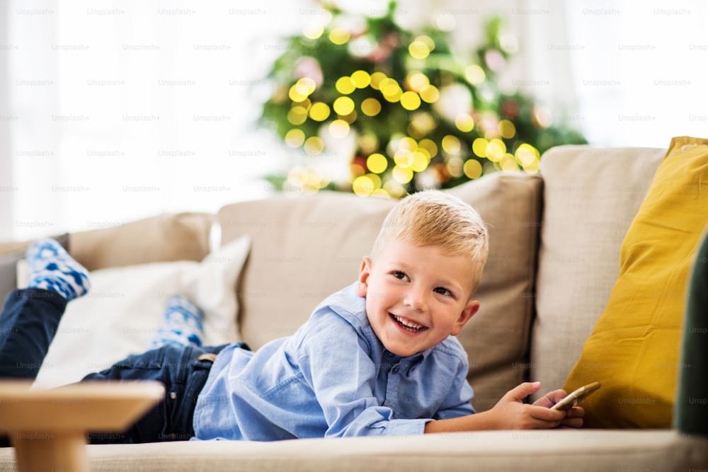 Ein kleiner Junge mit Smartphone, der zur Weihnachtszeit zu Hause auf einem Sofa liegt und Spiele spielt.