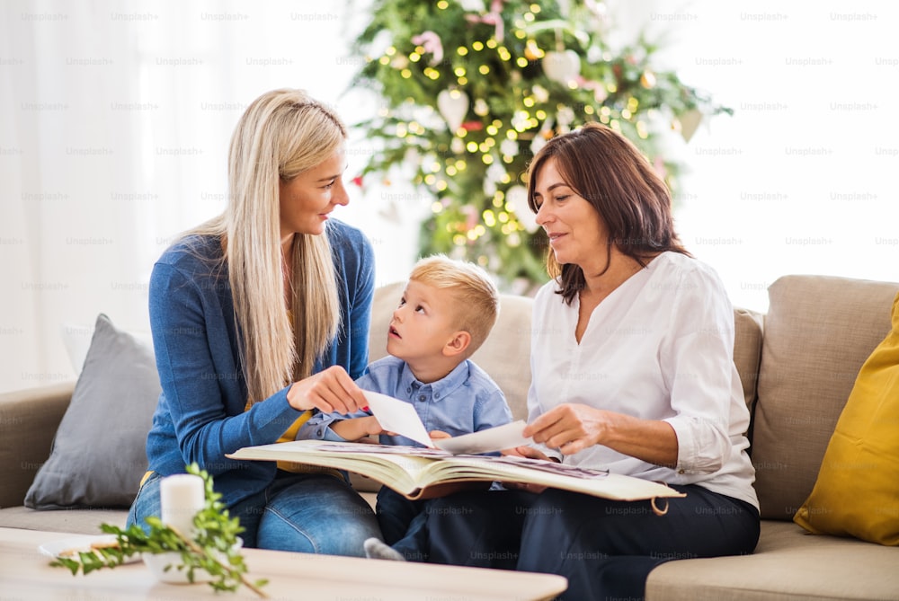 Um menino pequeno com mãe e avó sentados no sofá em casa na época do Natal, olhando fotos.