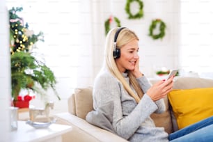 A young woman with headphones and smartphone sitting on a sofa at home at Christmas time, listening to music.