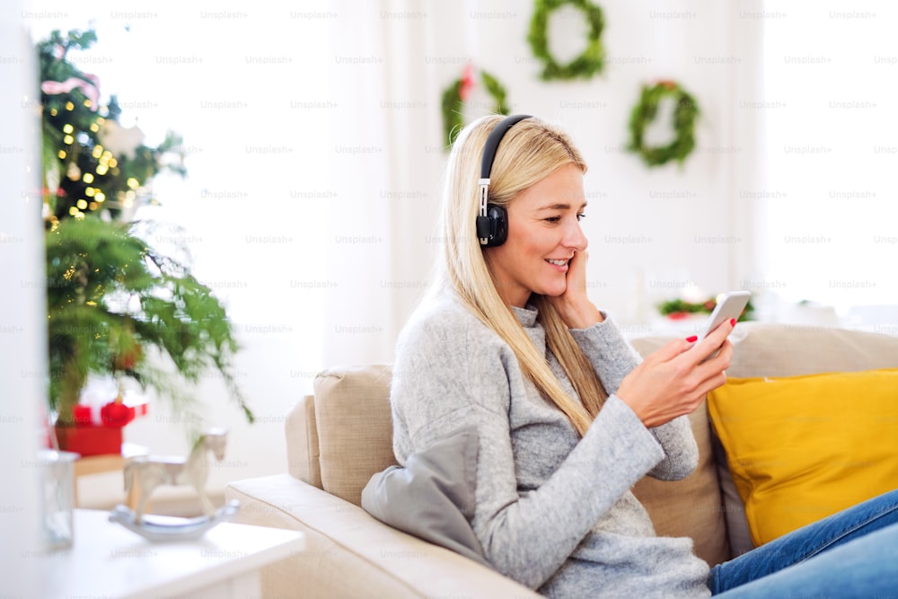 Una giovane donna con cuffie e smartphone seduta su un divano di casa nel periodo natalizio, ascoltando musica.