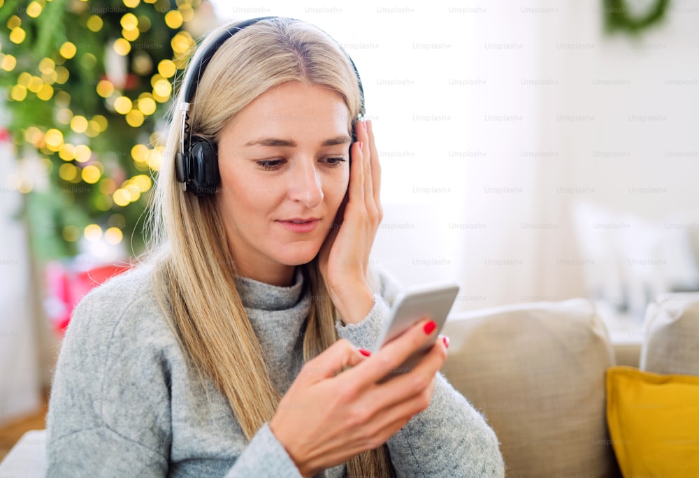 Uma jovem com fones de ouvido e smartphone sentada em um sofá em casa na época do Natal, ouvindo música.