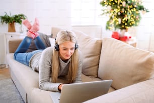 A young woman with headphones and laptop lying on a sofa at home at Christmas time.