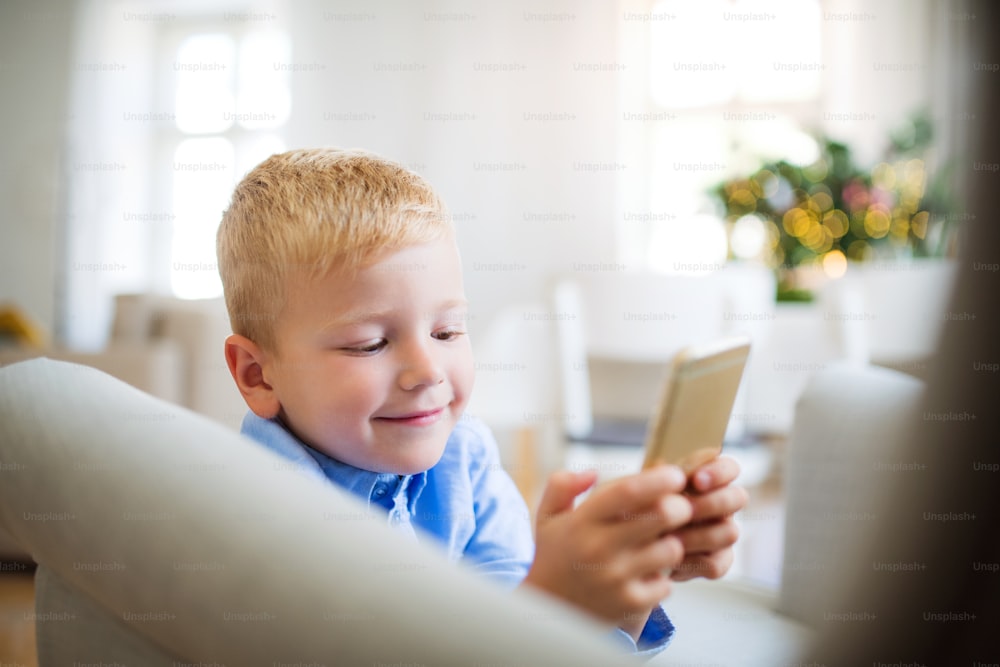 스마트폰을 들고 있는 어린 소년이 크리스마스 시간에 집에서 안락의자에 앉아 게임을 하고 있다.