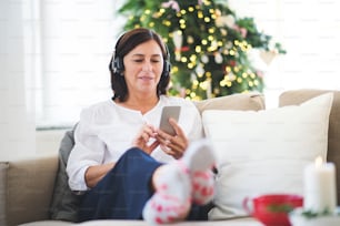 Una donna anziana con cuffie e smartphone seduta su un divano di casa, ascoltando musica nel periodo natalizio.
