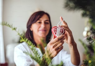 Uma mulher sênior segurando um enfeite em forma de coração, decorando uma árvore de Natal em casa.