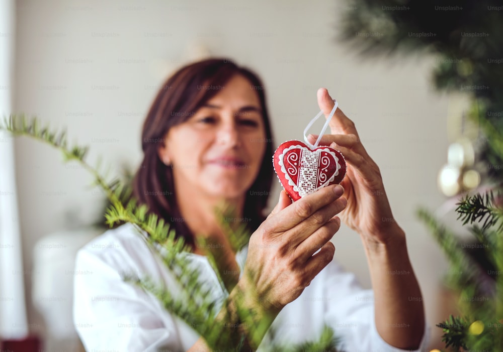 Una mujer mayor sosteniendo un adorno en forma de corazón, decorando un árbol de Navidad en casa.
