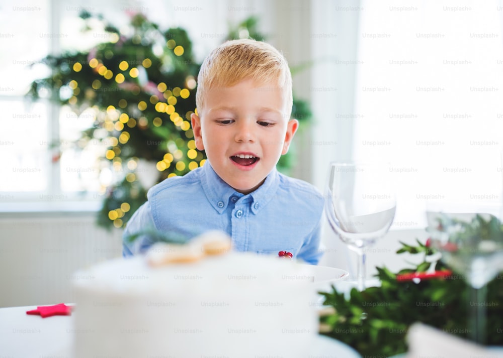 A small boy standing at the table at home at Christmas time, looking at a cake.