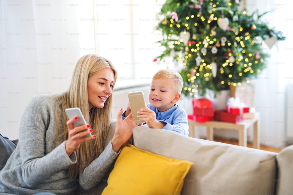 A mother and small boy with smartphones sitting on a sofa at home at Christmas time, playing games.