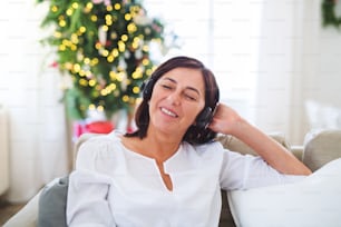 Uma mulher idosa com fones de ouvido sentada em um sofá em casa, ouvindo música na época do Natal.