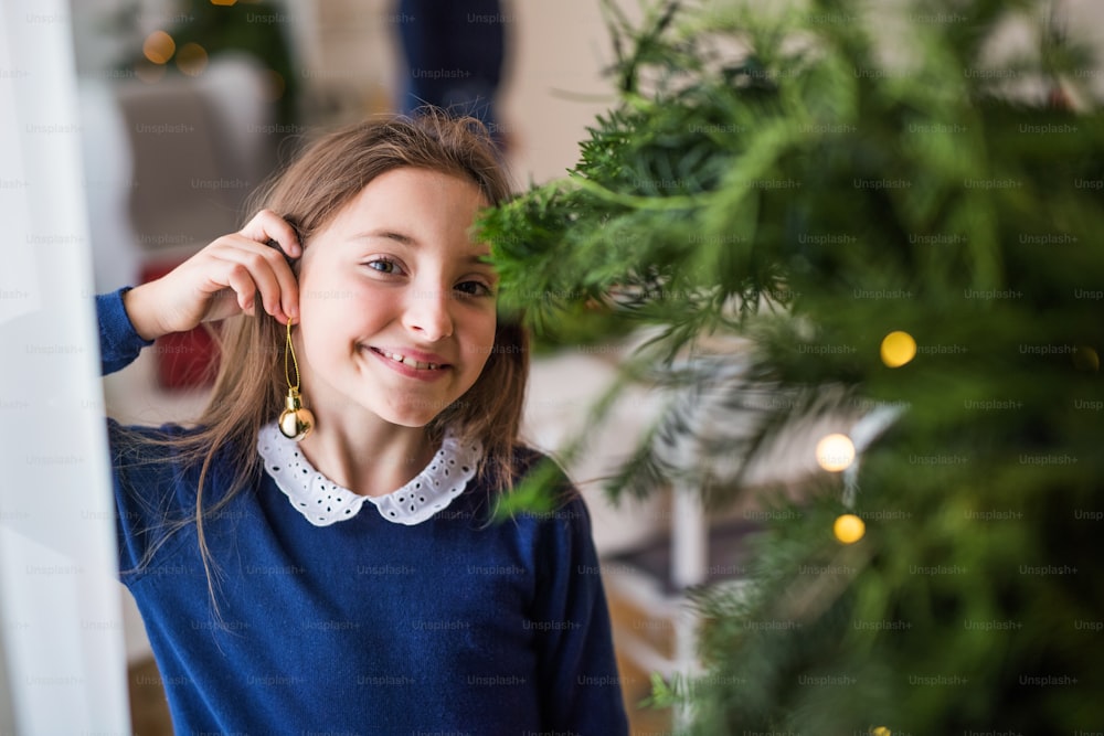 Small girl standing by a Christmas tree at home, putting a ball ornament on her ear as an earring. Copy space.