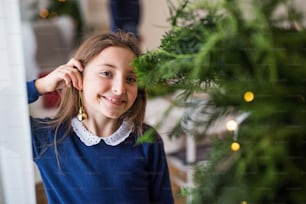 Bambina in piedi vicino a un albero di Natale a casa, mettendo un ornamento a sfera sul suo orecchino come orecchino. Copia spazio.