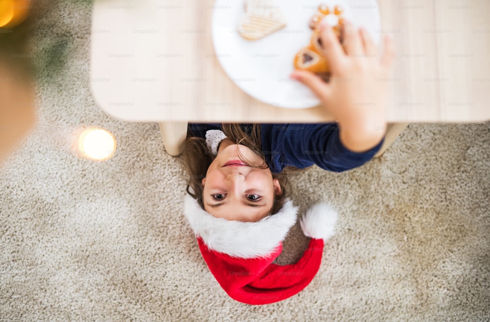 Une vue de dessus d’une petite fille avec un chapeau de Père Noël allongée sur le sol au moment de Noël, attrapant un biscuit.
