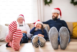 Una bambina con padre e nonno anziano seduti su un divano nel periodo natalizio, piedi su un tavolo e cappelli di Babbo Natale in testa.