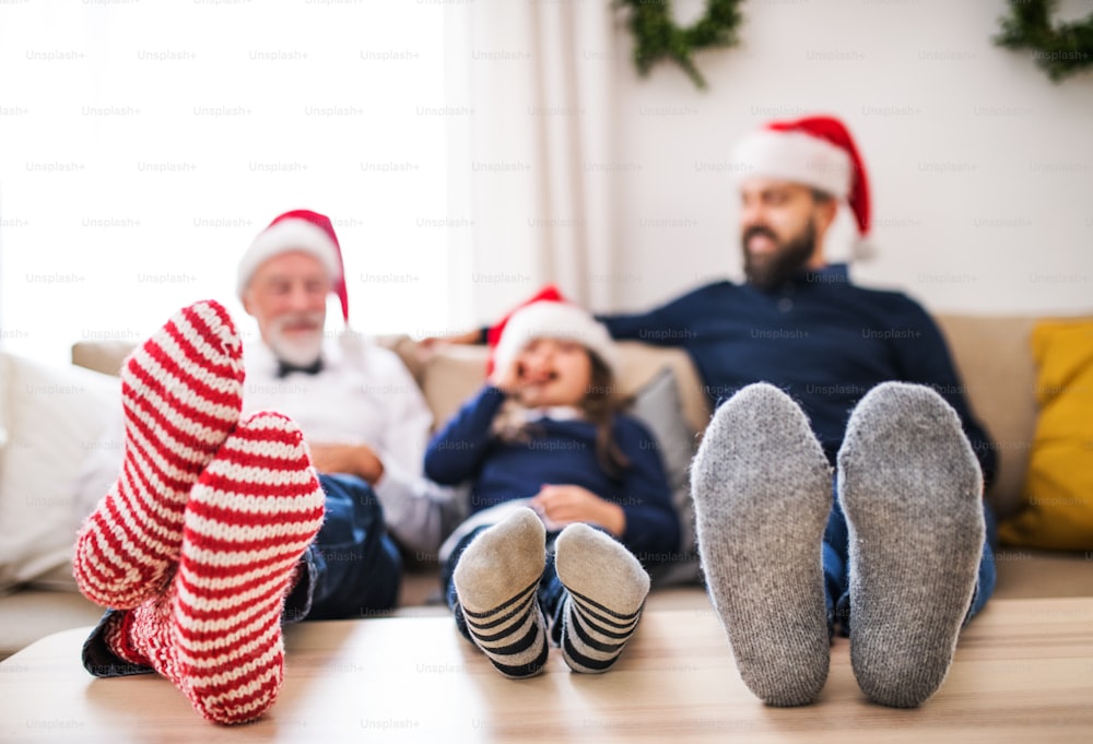 A small girl with father and senior grandfather sitting on a sofa at Christmas time, feet on a table and Santa hats on their head.