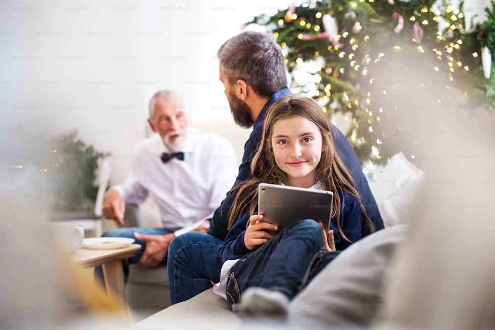 A small girl with tablet and her father and grandfather sitting on a sofa at Christmas time.