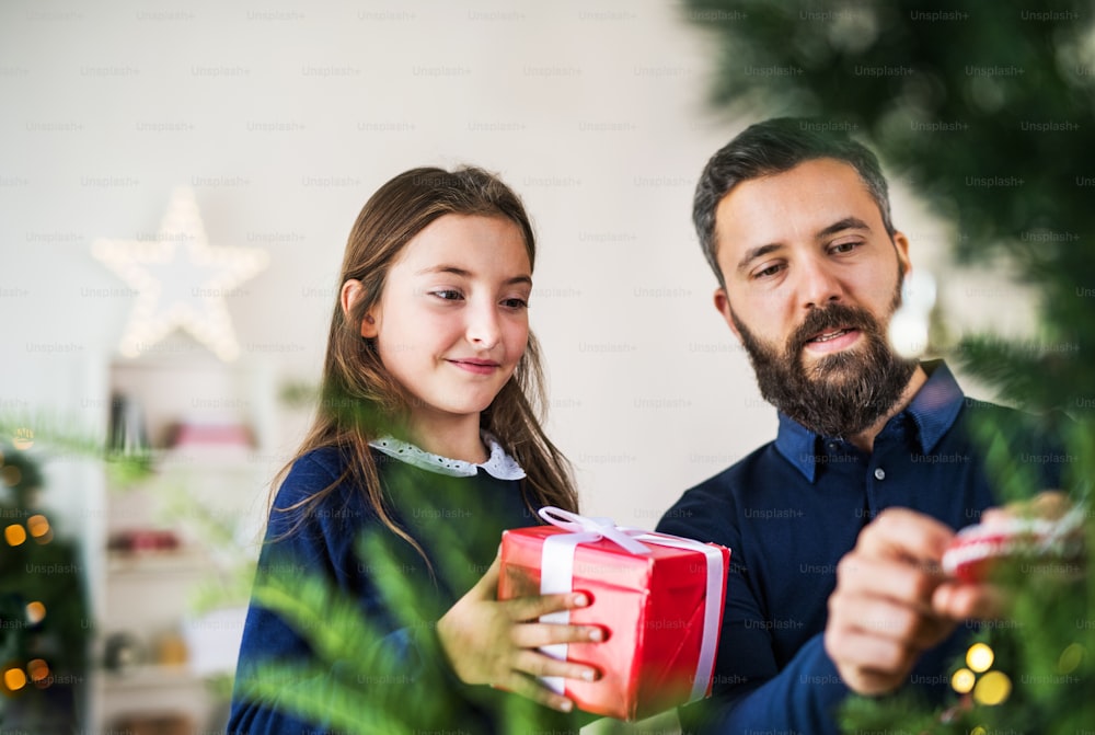 Una bambina con un regalo in piedi vicino a un albero di Natale con suo padre a casa.